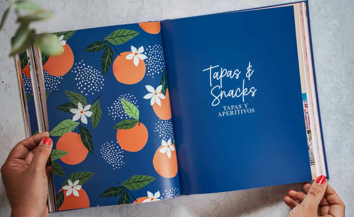 Die schönsten Kochbücher für den Sommer: Das Kochbuch Palma de Mallorca von Svenja Mattner-Shahi und Britta Welzer, erschienen im EMF Verlag. Das Bild zeigt eine Doppelseite aus dem Buch in Dunkelblau mit bunten Illustrationen von Orangen mit weißen Blüten und grünen Blättern sowie dem geletterten Kapitelaufmacher-Text Tapas und Snacks in Weiß