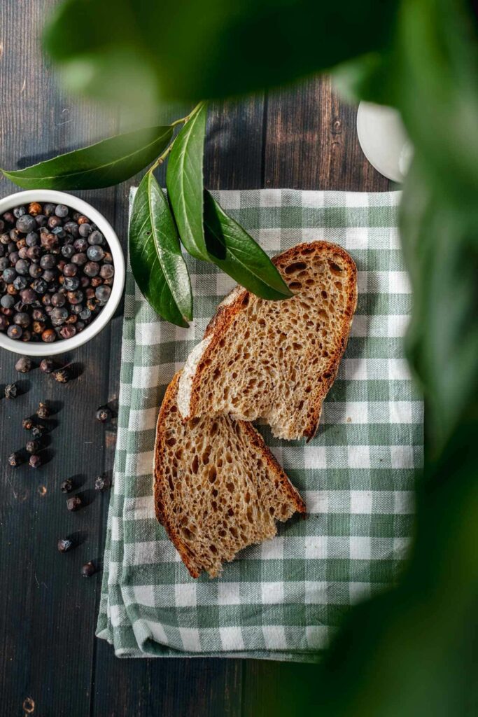 Rezept für fränkische Blaue Zipfel: Auf dem Bild ist eine grün-weiß-kariere Serviette, auf der zwei Scheiben Bauernbrot liegen sowie ein Schälchen mit Wacholder, aus dem einige Beeren auf die Tischplatte aus dunklem Holt gefallen sind. Im Vordergrund sind frische Lorbeerblätter zu sehen