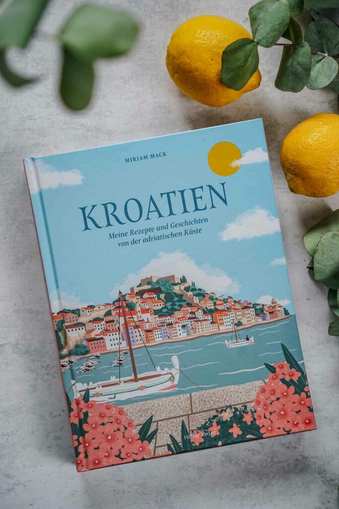 Die schönsten Kochbücher für den Sommer: Das Kroatien-Kochbuch Kroatien –Meine Rezepte und Geschichten von Miriam Mack aus dem Hölker-Verlag. Das Bild zeigt das Buchcover mit dem Titel in Dunkelblau auf einer illustrierten Szene der adriatischen Küste mit einem Segelboot auf dem Meer und einer Stadt im Hintergrund, umgeben von Eukalyptus-Blättern und Zitronen