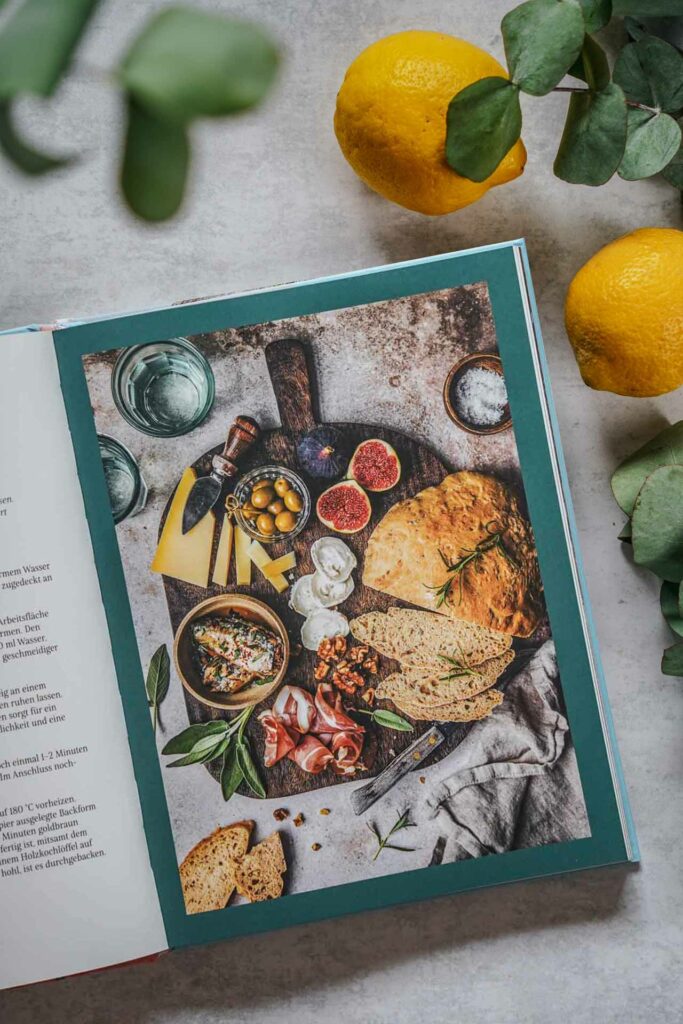 Die schönsten Kochbücher für den Sommer: Das Kroatien-Kochbuch Kroatien –Meine Rezepte und Geschichten von Miriam Mack aus dem Hölker-Verlag. Das Bild zeigt das für Aromabrot, das auf einem runden Holzbrett serviert wird mit Wurst- und Käsespezialitäten, Feigen und Oliven, umgeben von Eukalyptus-Blättern und Zitronen