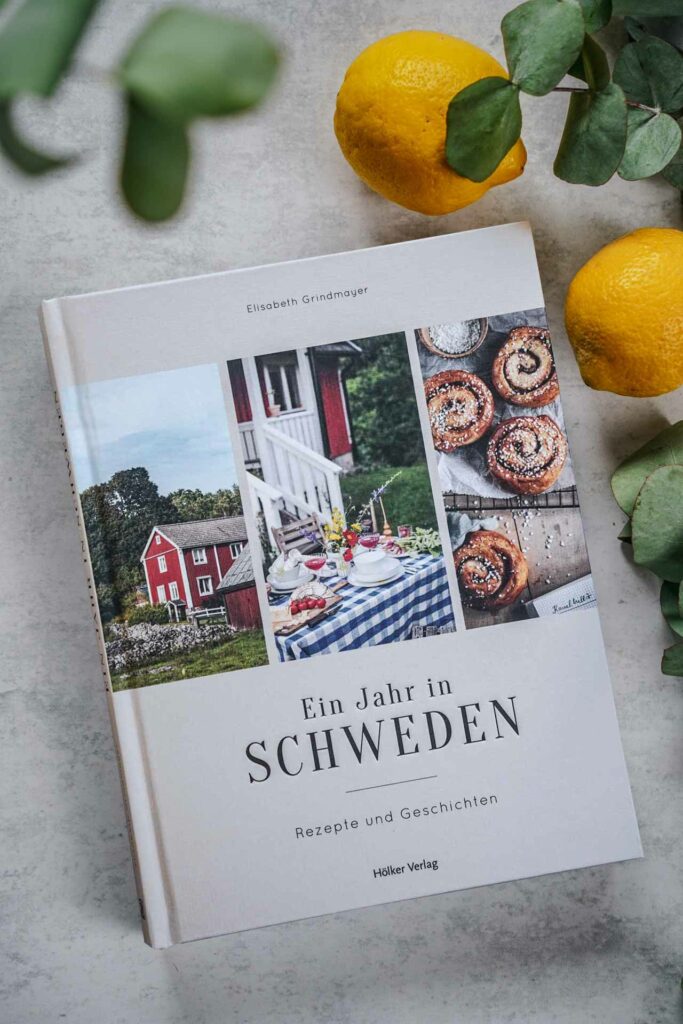 Die schönsten Kochbücher für den Sommer: Das Schweden-Kochbuch Ein Jahr in Schweden von Elisabeth Grindmayer aus dem Hölker-Verlag. Das Bild zeigt das Buchcover mit dem Titel in Schwarz auf beigefarbenem Leinen, umgeben von Eukalyptus-Blättern und Zitronen. Über dem Titel sind drei Fotos mit Impressionen aus Schweden: Links ein Schwedenhaus in Rot, in der Mitte die Veranda mit Garten mit einem sommerlich gedeckten Tisch mit blau karierter Tischdecke, Sommerblumen und Speisen, links frisch gebackene Zimtschnecken auf einem schwarzen Gitter auf einem Holztisch