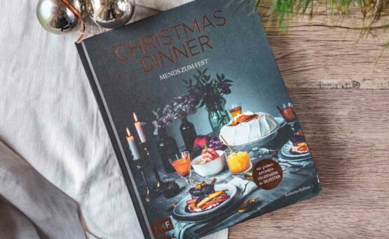 Die schönsten Kochbücher zu Weihnachten: Das Kochbuch Christmas Dinner, Menüs zum Fest, von Katharina Küllmer, erschienen im EMF Verlag. Das Cover in dunklem Petrol zeigt einen festlich gedeckten Tisch mit einem üppigen Menü mit Speisen und Getränken mit leuchtenden Farben in Wei0, Orange, Gelb und Rot, Kerzen und Blumen. Der Titel und das Logo schimmern in kupferfarbener Folie