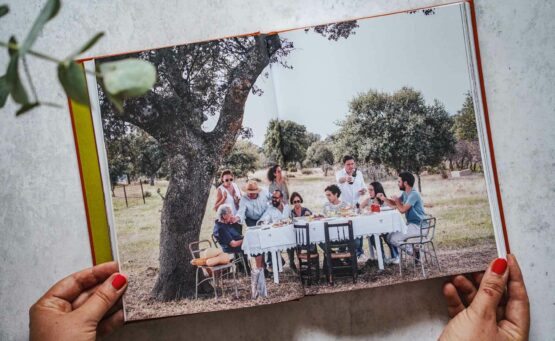 Die schönsten Kochbücher für den Sommer: Das Kochbuch Die spanische Landküche von José Pizarro, erschienen im Dorling Kindersley Verlag. Das Bild zeigt eine Doppelseite aus dem Buch, auf der der Autor mit Familie und Freunden an einem Tisch mit weißer Tischdecke draußen beim Essen ist, im Hintergrund ist eine typisch spanische Landschaft mit Olivenbäumen zu sehen