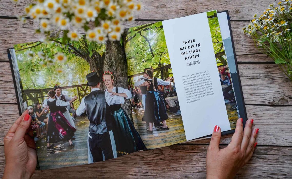 Die besten bayerischen Kochbücher: Zu Gast in Bayern, erschienen im Callwey Verlag. Das Bild zeigt tanzende Paare in traditioneller fränkischer Tracht auf der Limmersdorfer Lindenkirchweih. Das Buch liegt auf einem Holztisch, umgeben von weißen Wildblumen