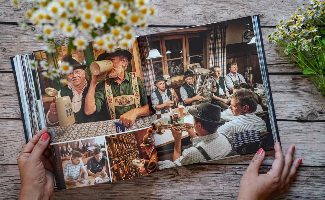 Die besten bayerischen Kochbücher: Zu Gast in Bayern, erschienen im Callwey Verlag. Das Bild zeigt Impressionen typisch bayerischer Musikantenstammtische in Tracht mit Blasmusik. Das Buch liegt auf einem Holztisch, umgeben von weißen Wildblumen