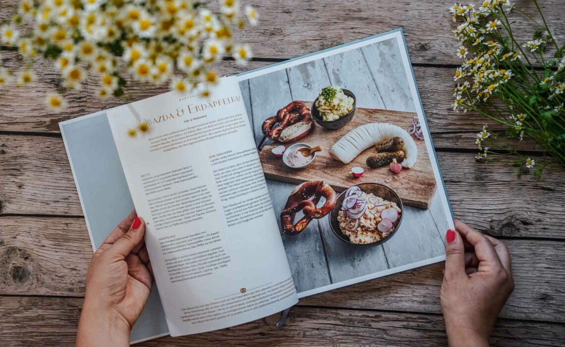 Die besten bayerischen Kochbücher: Dahoam von Alexander Huber,, erschienen im Dorling Kindersley Verlag. Das Bild zeigt das Rezept für eine Bayerische Brotzeit mit Obazda und Erdäpfelkas. Das Buch liegt auf einem Holztisch, umgeben von weißen Wildblumen