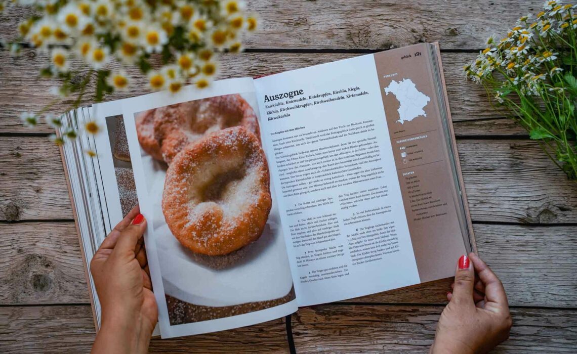 Die besten bayerischen Kochbücher: Das kulinarische Erbe Bayerns von Marion Reinhardt, erschienen im ars vivendi Verlag. Das Bild zeigt das Rezept für Auszogne. Das Buch liegt auf einem Holztisch, umgeben von weißen Wildblumen