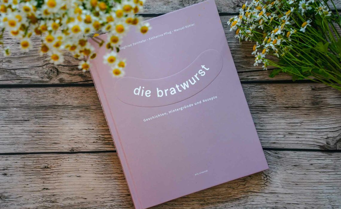 Kochbuch Die Bratwurst aus dem ars vivendi Verlag. Das Bild zeigt das Cover in Hellrosa mit dem Titel Die Bratwurst in Weiß darauf, der geschwungen in Form einer Bratwurst darauf abgebildet ist. Das Buch liegt auf einem Holztisch, umgeben von weißen Wildblumen