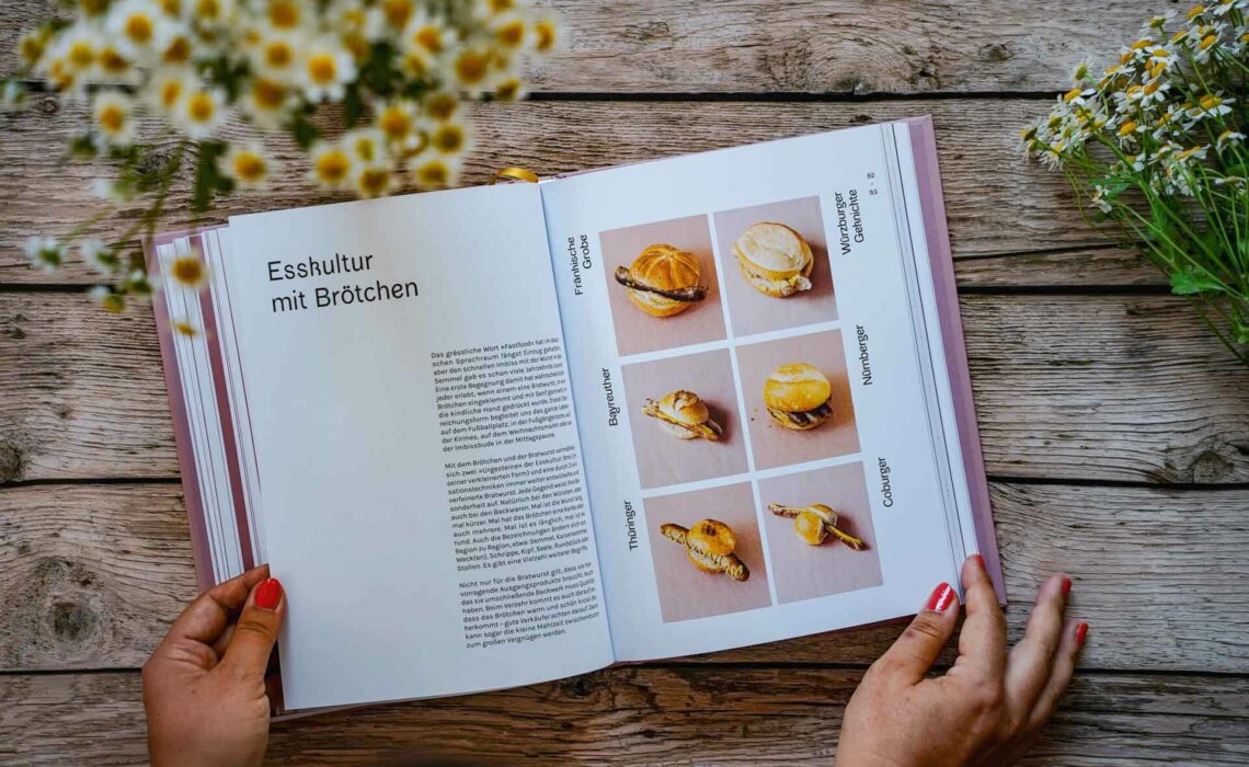 Das Kochbuch Die Bratwurst aus dem ars vivendi Verlag. Das Bild zeigt verschiedene Bratwürste im Brötchen. Das Buch liegt auf einem Holztisch, umgeben von weißen Wildblumen