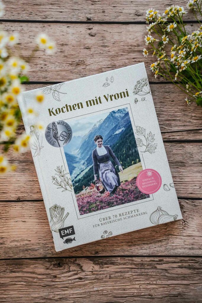 Die besten bayerischen Kochbücher: Einfach. Gut. Kochen mit Vroni von Vroni Lutz, erschienen im EMF Verlag. Das Bild zeigt die Autorin im Dirndl inmitten einer bayerischen Alpenlandschaft. Das Buch liegt auf einem Holztisch, umgeben von weißen Wildblumen