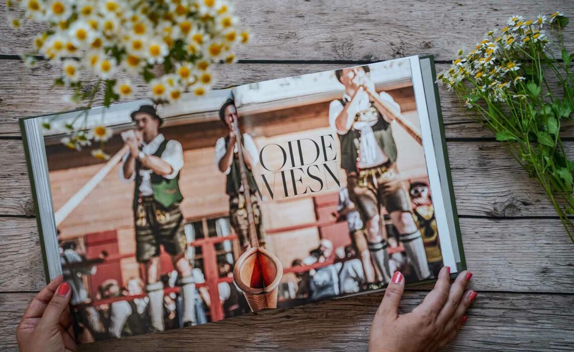 Die besten bayerischen Kochbücher: Zu Gast auf dem Oktoberfest, erschienen im Callwey Verlag. Das Bild zeigt den Kapitelaufmacher für die Oide Wiesn mit typisch bayerischen Blasmusikanten. Das Buch liegt auf einem Holztisch, umgeben von weißen Wildblumen