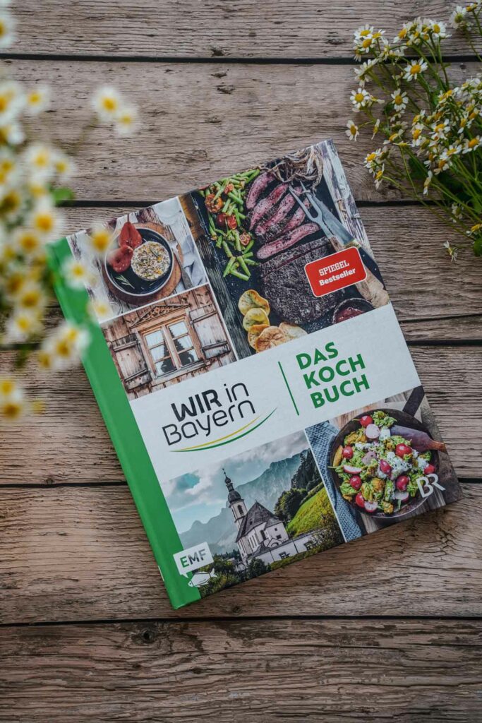 Die besten bayerischen Kochbücher: Wir in Bayern – Das Kochbuch, erschienen im EMF Verlag. Das Bild zeigt das Cover mit fünf Bildern, die Rezepte aus dem Kochbuch und bayerische Landschaften zeigen. Der Titel in Grün sowie das Logo Wir in Bayern stehen auf einem weißen Banner in der Mitte. Das Buch liegt auf einem Holztisch, umgeben von weißen Wildblumen