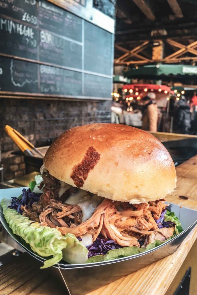 Weihnachten in London: Der Borough Market ist ein echtes Foodie-Paradies, auf dem es sehr viel Street Food gibt. Auf dem Bild ist ein Pulled Pork Burger in einer schwarzen to-go-Schale zu sehen, im Hintergrund eine große Angebotstafel mit bunter Kreideschrift an der Wand