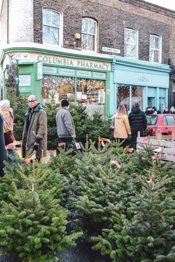 Weihnachten in London: Weihnachtsbäume zum Kauf auf dem berühmten Columbia Road Flower Market mit typischen Backsteinhäusern und bunten Ladenfassaden im Hintergrund
