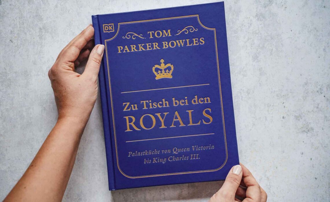 Das Kochbuch Zu Tisch bei den Royals – Palastküche von Queen Victoria bis King Charles III. von Tom Parker Bowles, erschienen im Dorling Kindersley Verlag. Das Bild zeigt das Cover in Royalblau. Der Titel ist mit Goldfolie in einem Rahmen darauf gedruckt