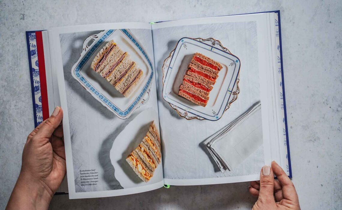 Das Kochbuch Zu Tisch bei den Royals – Palastküche von Queen Victoria bis King Charles III. von Tom Parker Bowles, erschienen im Dorling Kindersley Verlag. Das Bild zeigt eine Doppelseite für das Rezept für Afternoon Tea Sandwiches mit verschiedenen Füllungen. Die Sandwiches sind auf verschiedenen Servierplatten drapiert, daneben liegt eine weiße Servieette