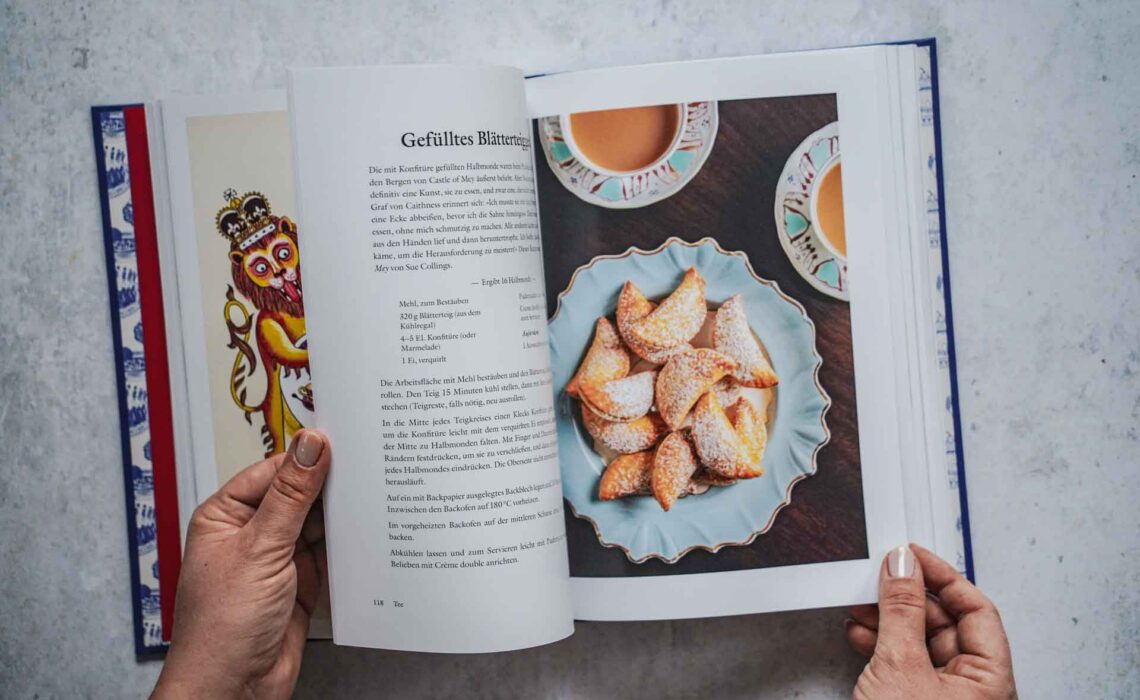 Das Kochbuch Zu Tisch bei den Royals – Palastküche von Queen Victoria bis King Charles III. von Tom Parker Bowles, erschienen im Dorling Kindersley Verlag. Das Bild zeigt das Rezept für gefülltes Blätterteiggebäck. Das halbmondförmige, mit Puderzucker bestäubte Gebäck liegt auf einem hellblauen Porzellanteller, umgeben von zwei Teetassen mit schwarzem Tee und Milch in blau gemustertem Porzellantassen