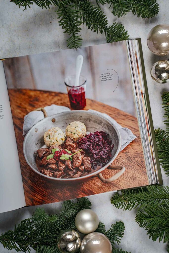 Kochbücher zu Weihnachten: Zu Gast auf dem Oktoberfest von Franz Kotteder, erschienen im Callwey Verlag. Das Bild zeigt das Rezept für Wildgulasch mit Blaukraut und Semmelknödeln. Das Buch liegt auf einem grauen Untergrund, umgeben von grünen Tannenzweigen und Weihnachtskugeln in Silber und Cremefarben