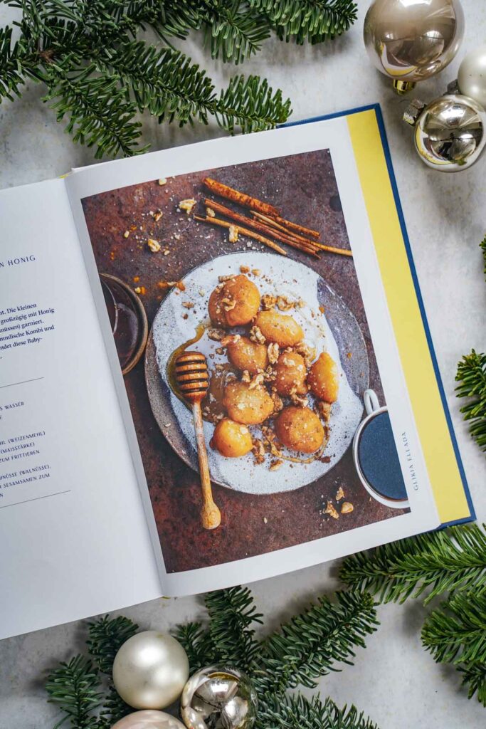 Kochbücher zu Weihnachten: Das Kochbuch Sonne und Meer auf dem Teller von Lazaros Kapageoroglou, erschienen im AT Verlag. Das Bild zeigt das Rezeptfoto für Loukoumades me Meli – Baby-Donuts, getränkt in Honig. Das Buch liegt auf einem grauen Untergrund, umgeben von grünen Tannenzweigen und Weihnachtskugeln in Silber und Cremefarben