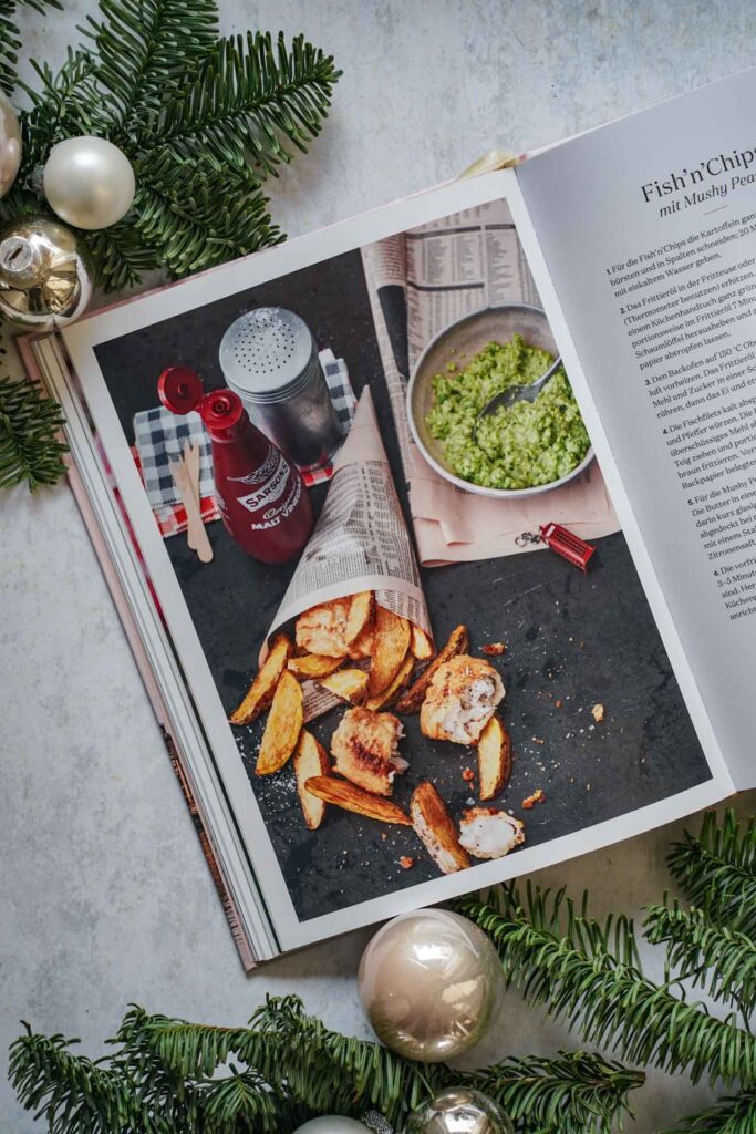 Kochbücher zu Weihnachten: Das England-Kochbuch London Love Story – Rezepte und Geschichten von Anne-Katrin Weber, erschienen im Hölker Verlag. Das Bild zeigt das Rezeptfoto für Fish and Chips mit Mushy Peas. Das Buch liegt auf einem grauen Untergrund, umgeben von grünen Tannenzweigen und Weihnachtskugeln in Silber und Cremefarben