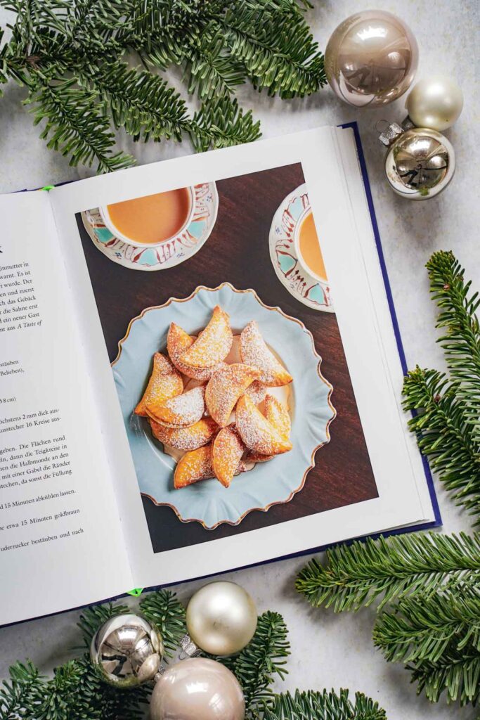 Kochbücher zu Weihnachten: Das Kochbuch Zu Tisch bei den Royals von Tom Parker Bowls, erschienen im Dorling Kindersley Verlag. Das Bild zeigt das Rezeptfoto für gefüllte Blätterteiggebäck mit Puderzucker bestreut. Das Buch liegt auf einem grauen Untergrund, umgeben von grünen Tannenzweigen und Weihnachtskugeln in Silber und Cremefarben