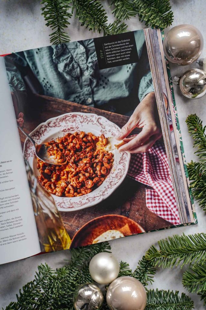 Kochbücher zu Weihnachten: Roma in Cucina von Deborah Ferrini-Kreitmair erschienen im EMF Verlag. Das Bild zeigt das Rezeptbild für Braune Bohnen mit Tomatensauce und rohem Schinken. Das Buch liegt auf einem grauen Untergrund, umgeben von grünen Tannenzweigen und Weihnachtskugeln in Silber und Cremefarben