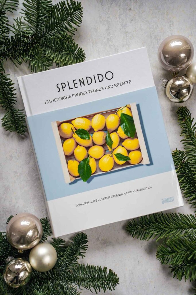 Kochbücher zu Weihnachten: Das Kochbuch Splendido. Italienische Produktkunde und Rezepte von Mercedes Lauenstein und Juri Gottschall, erschienen im DuMont Buchverlag Das Bild zeigt das Cover in Weiß mit einer hellblauen Banderole, auf der eine weiße Kiste mit Zitronen abgebildet ist. Das Buch liegt auf einem grauen Untergrund, umgeben von grünen Tannenzweigen und Weihnachtskugeln in Silber und Cremefarben