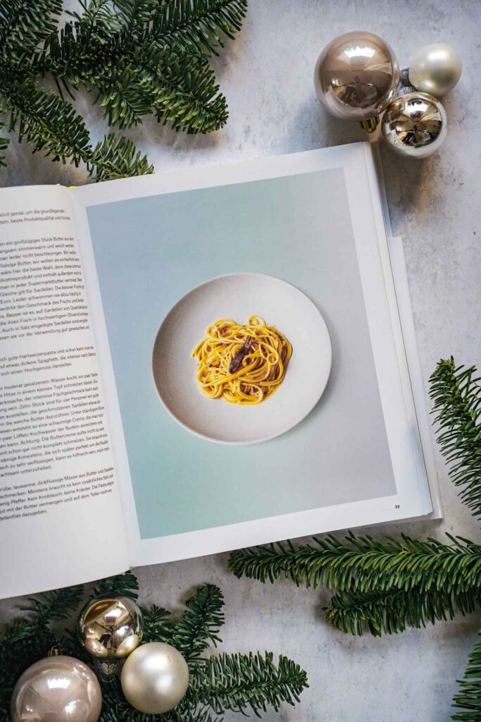 Kochbücher zu Weihnachten: Das Kochbuch Splendido. Italienische Produktkunde und Rezepte von Mercedes Lauenstein und Juri Gottschall, erschienen im DuMont Buchverlag Das Bild zeigt das Rezeptoto für Spaghetti mit Butter und Sardellen. Das Buch liegt auf einem grauen Untergrund, umgeben von grünen Tannenzweigen und Weihnachtskugeln in Silber und Cremefarben