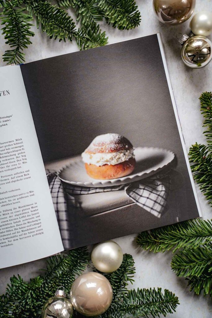 Kochbücher zu Weihnachten: Das Schweden-Kochbuch Ein Jahr in Schweden von Elisabeth Grindmayer aus dem Hölker Verlag. Das Bild zeigt das Rezeptfoto für Semlor – Schwedische Mandelkrapfen. Das Buch liegt auf einem grauen Untergrund, umgeben von grünen Tannenzweigen und Weihnachtskugeln in Silber und Cremefarben