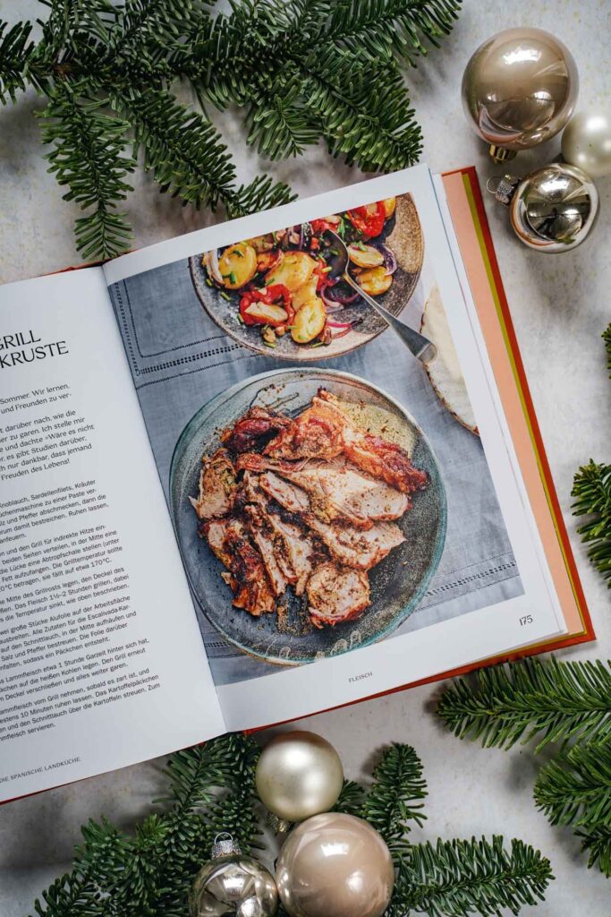 Kochbücher zu Weihnachten: Das Kochbuch Die spanische Landküche von José Pizarro, erschienen im Dorling Kindersley Verlag. Das Bild zeigt das Rezeptfoto für Lammschulter vom Grill mit Oliven-Kräuter-Kruste. Das Buch liegt auf einem grauen Untergrund, umgeben von grünen Tannenzweigen und Weihnachtskugeln in Silber und Cremefarben