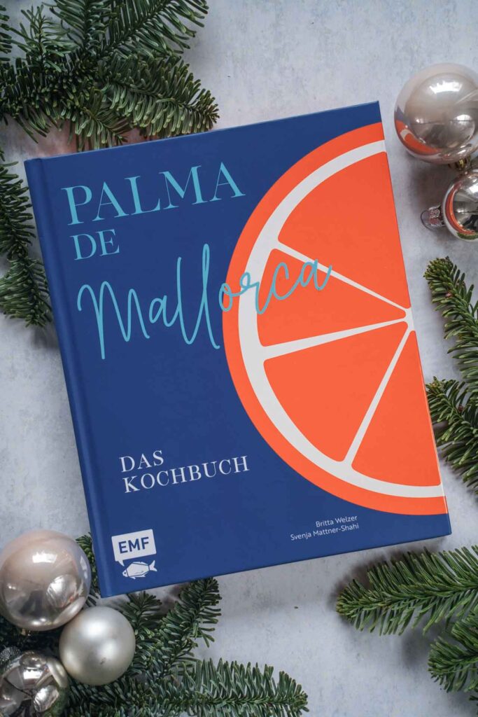 Kochbücher zu Weihnachten: Das Kochbuch Palma de Mallorca von Svenja Mattner-Shahi und Britta Welzer, erschienen im EMF Verlag. Das Bild zeigt das Buchcover in Dunkelblau und einer illustrierten Orange, der Buchtitel in Hellblau darauf. Das Buch liegt auf einem grauen Untergrund, umgeben von grünen Tannenzweigen und Weihnachtskugeln in Silber und Cremefarben