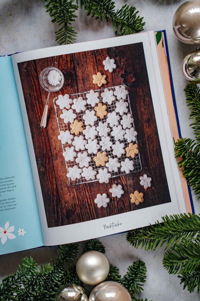 Kochbücher zu Weihnachten: Das Kochbuch Palma de Mallorca von Svenja Mattner-Shahi und Britta Welzer, erschienen im EMF Verlag. Das Bild zeigt das Rezeptbild für Crespells, einfache Kekse mit Puderzucker bestreut. Das Buch liegt auf einem grauen Untergrund, umgeben von grünen Tannenzweigen und Weihnachtskugeln in Silber und Cremefarben