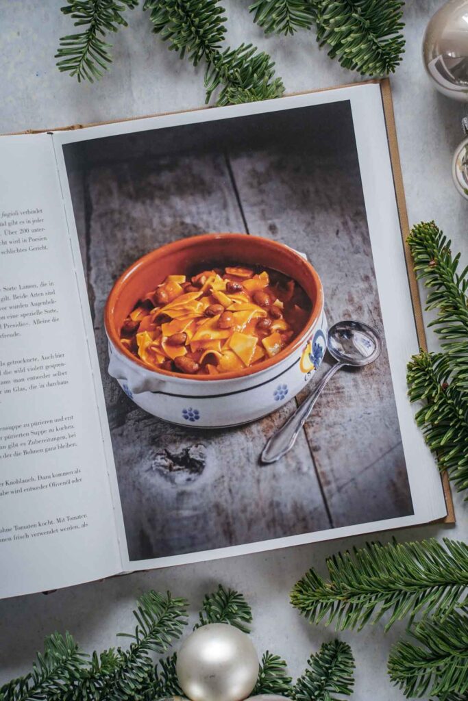 Kochbücher zu Weihnachten: Das Italien-Kochbuch alla buona von Claudio del Principe, erschienen im AT Verlag. Das Bild zeigt das Rezeptfoto für Pasta e Fagioli, Nudeln mit Bohnen. Das Buch liegt auf einem grauen Untergrund, umgeben von grünen Tannenzweigen und Weihnachtskugeln in Silber und Cremefarben
