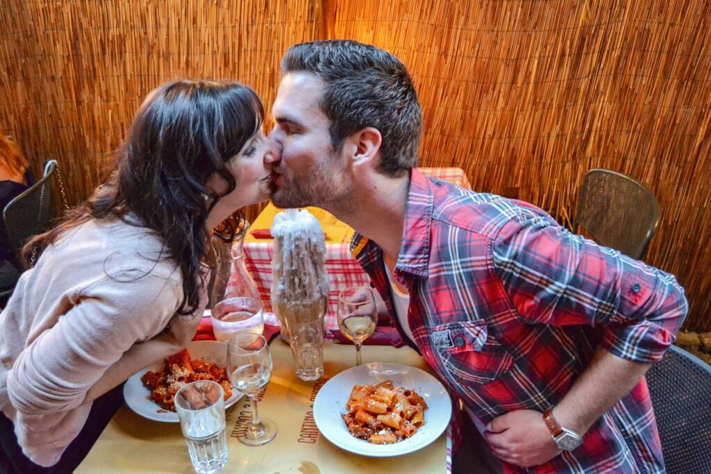 Alexandra und Florian von Tables & Fables in einer Hosteria in Rom. Sie küssen sich über zwei Teller mit Pasta all'Amatriciana hinweg