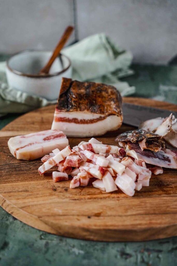 Originalrezept für Pasta all'Amatriciana: Ein Holzbrett aus Akazie mit einem Stück Guanciale, im Vordergrund sind abgeschnittene Guanciale-Streifen zu sehen, im Hintergrund eine kleine Schale mit gehobeltem Pecorino Romano und einem Holzlöffel und eine mintfarbene Serviette