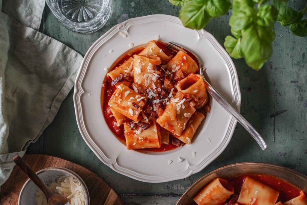 Originalrezept für Pasta all'Amatriciana: ein Teller mit Pacchheri in Tomatensauce mit knusprigem Guanciale und gehobeltem Pecorino Romano auf einem grünen Holztisch, umgeben von Basilikum, einer mintfarbenem Serviette, einem Glas Wasser und gehobeltem Pecorino in einer Schale