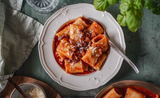 Originalrezept für Pasta all'Amatriciana: ein Teller mit Pacchheri in Tomatensauce mit knusprigem Guanciale und gehobeltem Pecorino Romano auf einem grünen Holztisch, umgeben von Basilikum, einer mintfarbenem Serviette, einem Glas Wasser und gehobeltem Pecorino in einer Schale