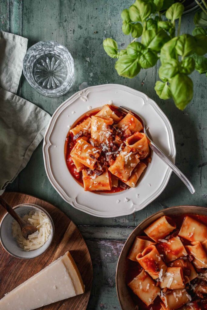 Rezept für Pasta all'Amatriciana: ein Teller mit Pacchheri in Tomatensauce mit knusprigem Guanciale und gehobeltem Pecorino Romano auf einem grünen Holztisch, umgeben von Basilikum, einer mintfarbenem Serviette, einem Glas Wasser, einem Stück Pecorino Romano auf einem Holzbrett und einer Pfanne mit mehr Pasta