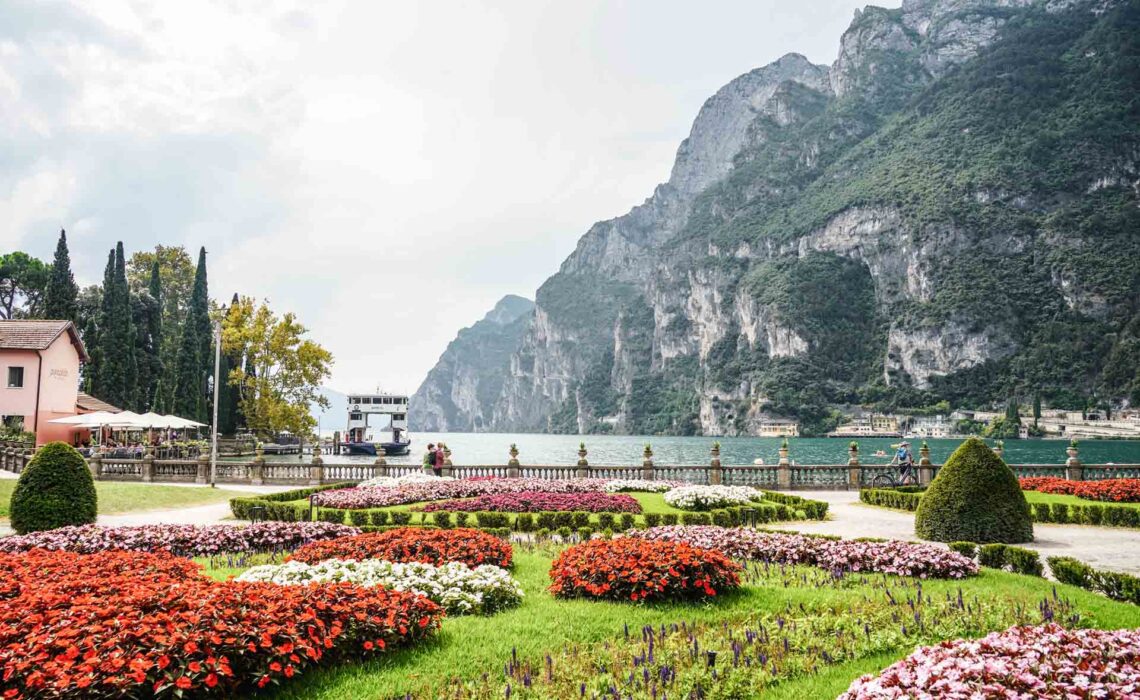Die schönsten Orte am Gardasee: die Giardini all'Italiana di Punta Lido in Riva del Garda mit einem bunten Blütenmeer und dem Gardasee und dem Bergpanorama im Hintergrund