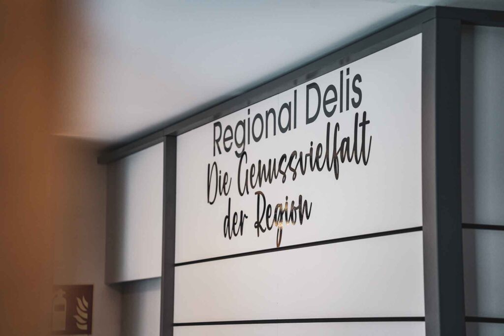 Schild in der Genussecke in der Tourist Info im Handwerkerhof Nürnberg mit schwarzer Schrift auf weißem Hintergrund: Regional Delis: Die Genussvielfalt der Region