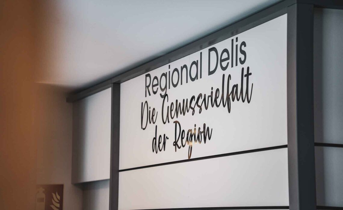 Schild in der Genussecke in der Tourist Info im Handwerkerhof Nürnberg mit schwarzer Schrift auf weißem Hintergrund: Regional Delis: Die Genussvielfalt der Region