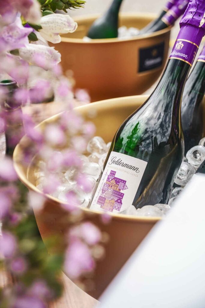 Geldermann x KPM –Launch der Bayreuther Festspiel-Cuvée zu Richard Wagners Die Meistersinger von Nürnberg 2025: Die Sektflasche mit Etikett mit stilisierten Fachwerkhäusern in Lila und Gold in einem goldenen Sektkühler mit Eiswürfeln