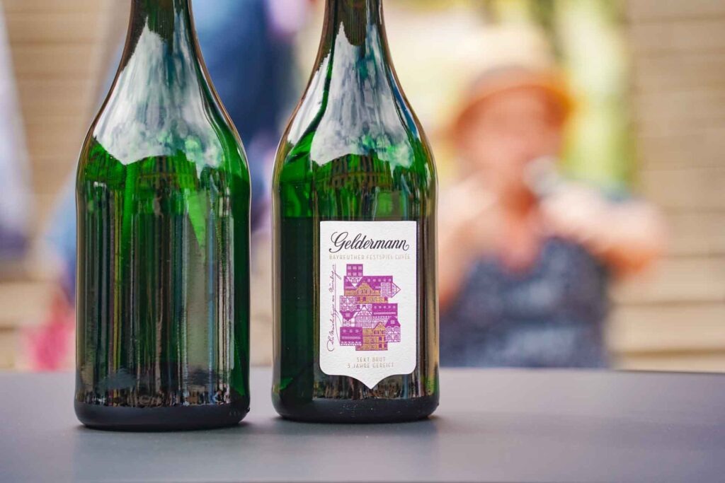 Geldermann x KPM –Launch der Bayreuther Festspiel-Cuvée zu Richard Wagners Die Meistersinger von Nürnberg 2025: Das Bild zeigt zwei Sektflaschen, eine davon von hinten, eine von vorne mit dem Etikett der Festspiel-Cuvée mit Fachwerkhäusern in Gold und Lila. Im Hintergrund unscharf ist Schauspieler Manni Laudenbach im Tannhäuser-Talk zu sehen