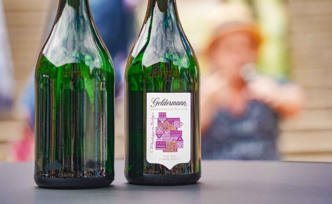 Geldermann x KPM –Launch der Bayreuther Festspiel-Cuvée zu Richard Wagners Die Meistersinger von Nürnberg 2025: Das Bild zeigt zwei Sektflaschen, eine davon von hinten, eine von vorne mit dem Etikett der Festspiel-Cuvée mit Fachwerkhäusern in Gold und Lila. Im Hintergrund unscharf ist Schauspieler Manni Laudenbach im Tannhäuser-Talk zu sehen