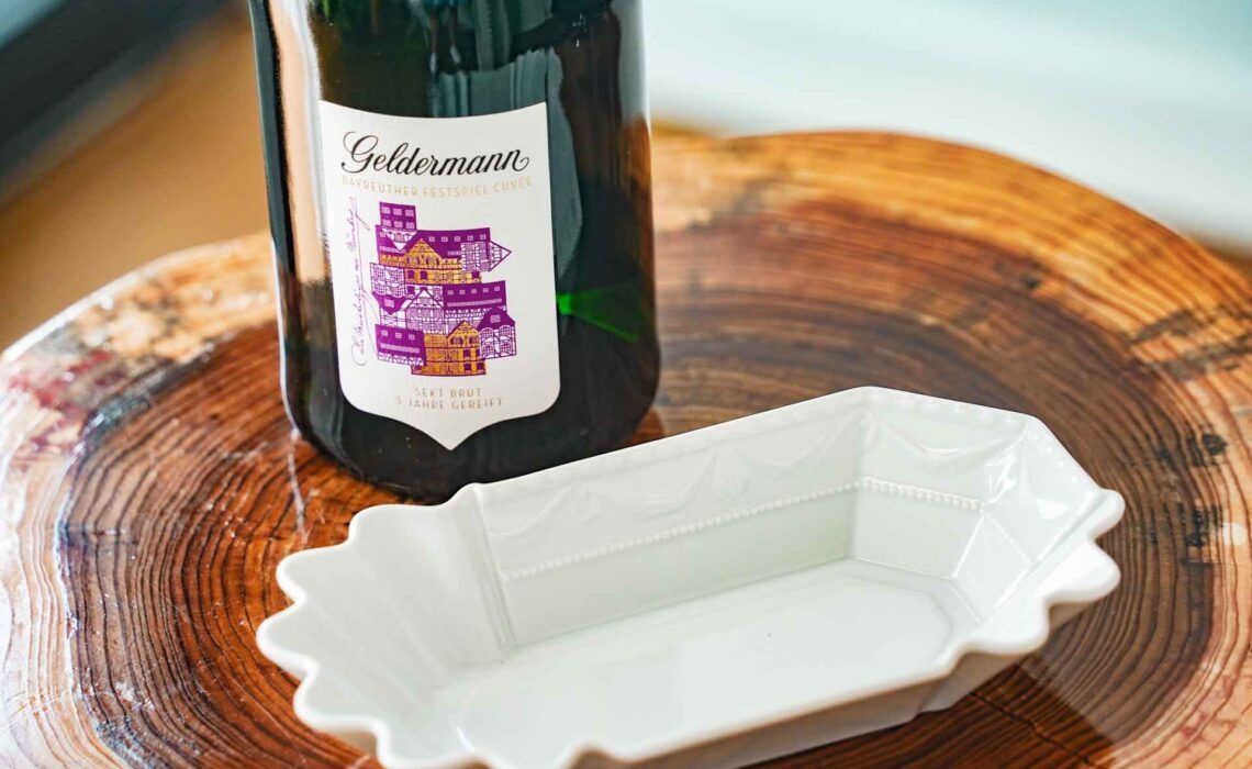 Geldermann x KPM –Launch der Bayreuther Festspiel-Cuvée zu Richard Wagners Die Meistersinger von Nürnberg 2025: Festspiel-Cuvée Flasche und KPM KURLAND Currywurstschale aus Porzellan auf einem Holztisch in Form eines Baumstumpfes