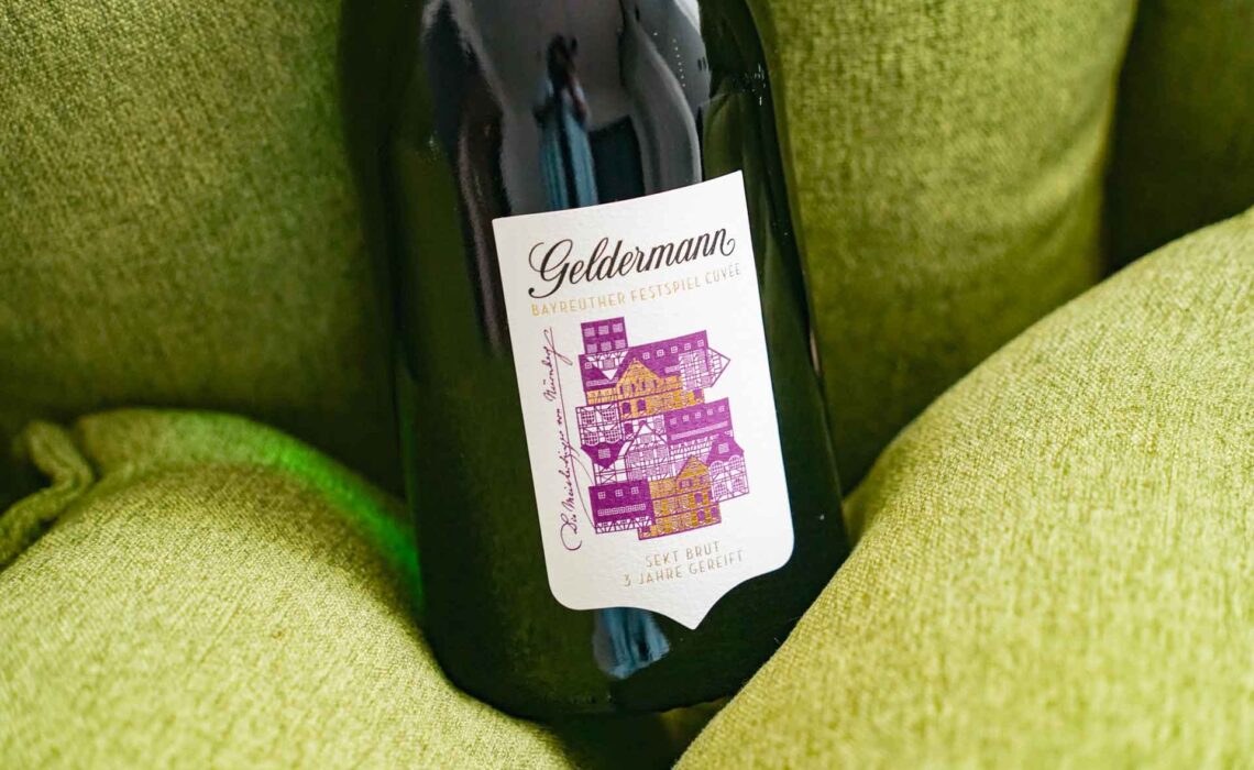 Geldermann x KPM –Launch der Bayreuther Festspiel-Cuvée zu Richard Wagners Die Meistersinger von Nürnberg 2025: Die Flasche mit dem Etikett. aus stilisierten Fachwerkhäusern in Lila und Gold auf dem grünen Designer-Sessel