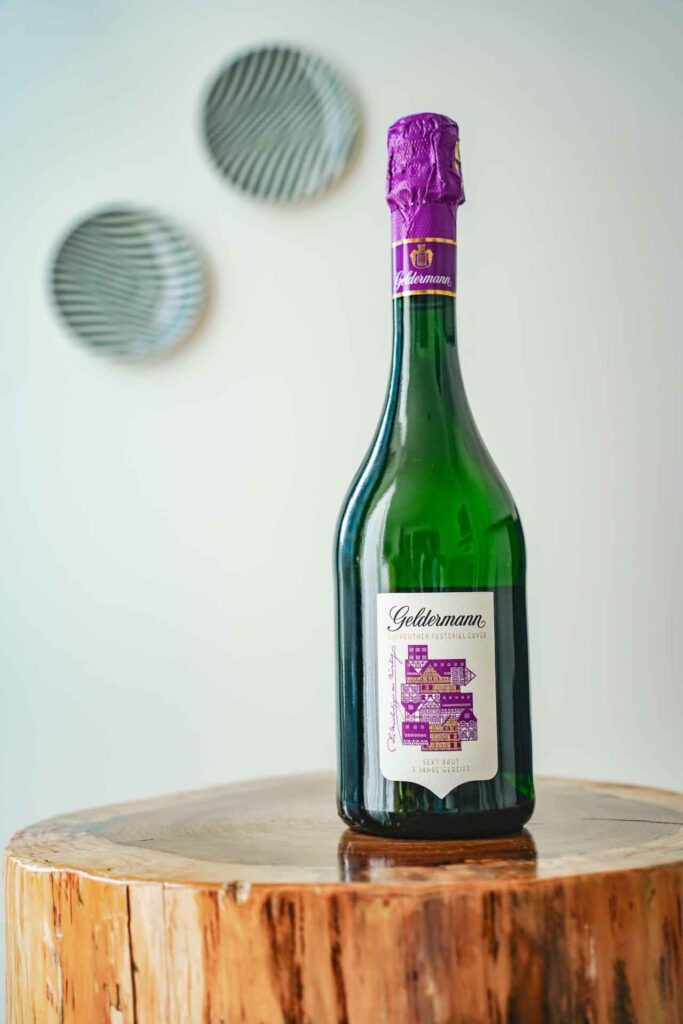 Geldermann x KPM –Launch der Bayreuther Festspiel-Cuvée zu Richard Wagners Die Meistersinger von Nürnberg 2025: Improvisiertes Fotoshooting im Designzimmer im KPM-Hotel mit der Geldermann Festspiel-Cuvée auf einem Holzstamm vor einer weißen Wand, im Hintergrund zwei Porzellanteller in Schwarz-Weiß