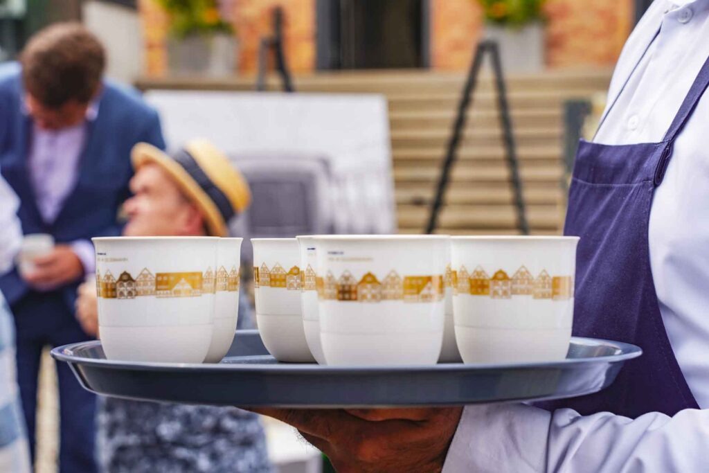 Geldermann x KPM –Launch der Bayreuther Festspiel-Cuvée zu Richard Wagners Die Meistersinger von Nürnberg 2025: KPM LAB Porzellanbecher mit gold illustrierten Fachwerkhäusern auf einem blauen Tablett
