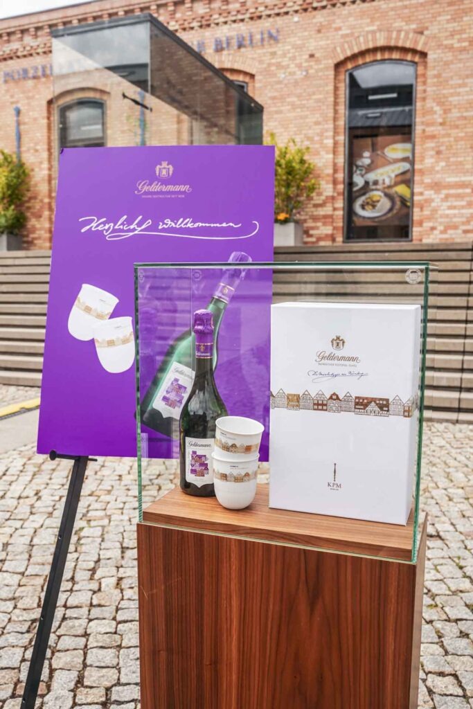 Geldermann x KPM –Launch der Bayreuther Festspiel-Cuvée zu Richard Wagners Die Meistersinger von Nürnberg 2025: Präsentation der Fespiel-Cuvée und der KPM-Becher im Set in einer Vitrine mit Holzsockel mit einem Plakataufsteller in Lila im Hintergrund