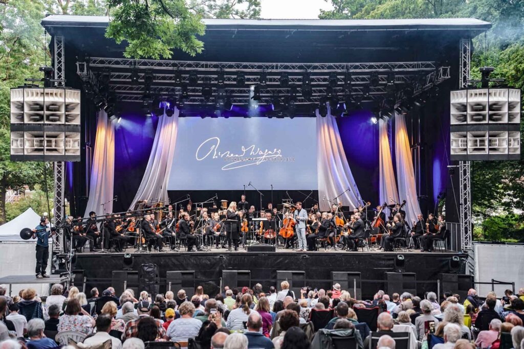 Mit Geldermann bei den Bayreuther Festspielen 2025: Festspiel-Open-Air im Festspielpark. Auf der Bühne ist das Festspielorchester zu sehen, davor Moderator Axel Brüggemann im Gespräch mit der Festspielchefin der Bayreuther Festspiele, Katharina Wagner. Im Vordergrund ist das Publikum im voll besetzten Festspielpark zu sehen